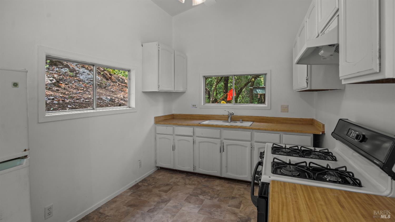 Vacant kitchen with white cabinets, wood countertops, and windows overlooking trees and a rocky slope.