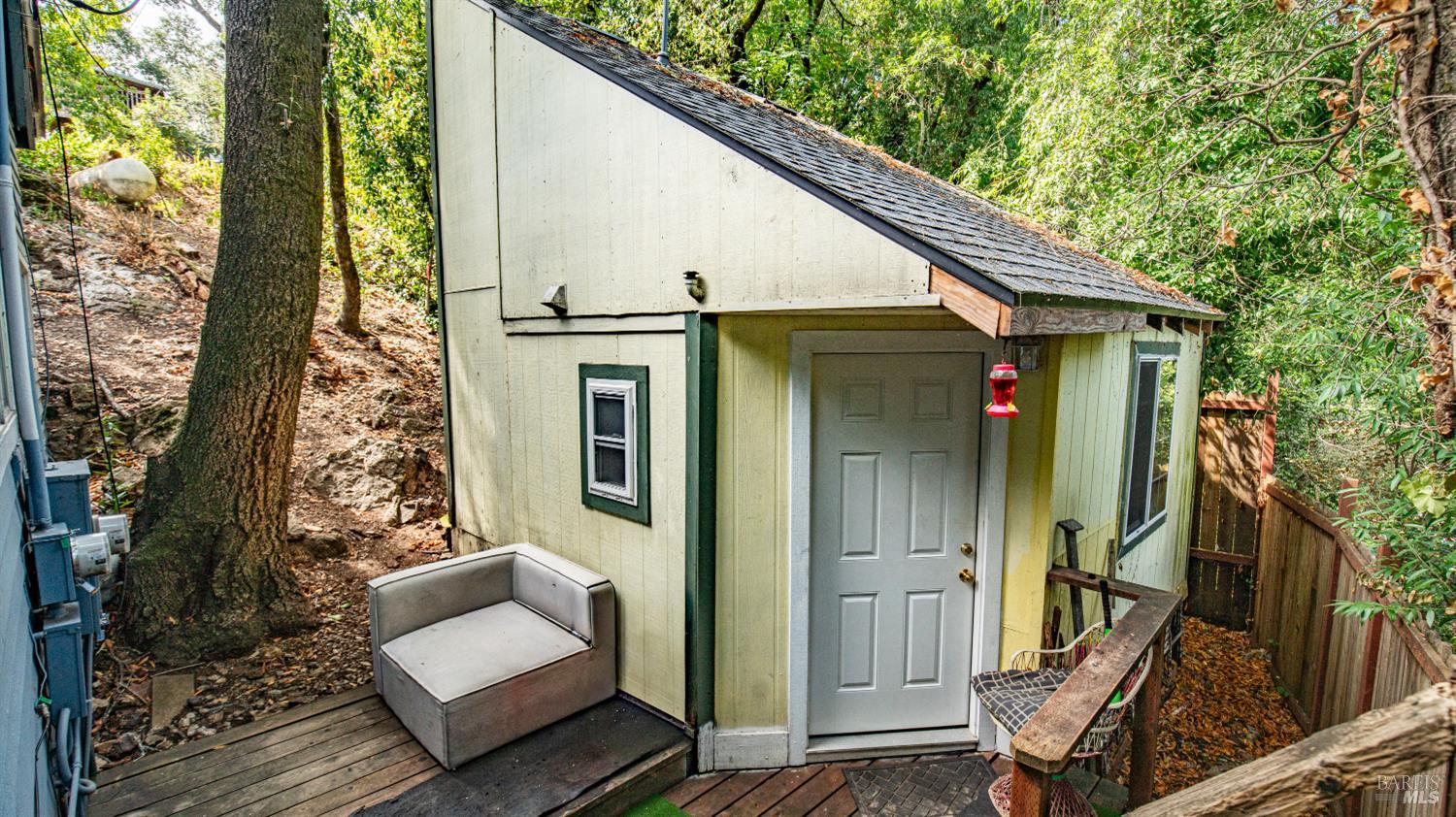 Small pale green outbuilding with a steep roof and wooden deck among trees.