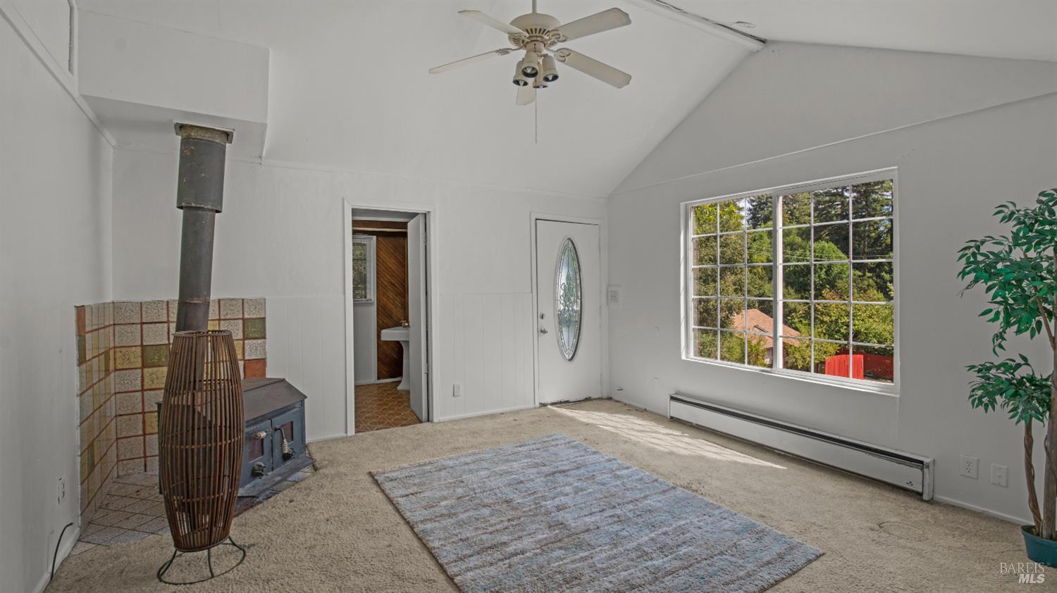 Living room with vaulted ceiling, wood-burning stove, and large window overlooking trees.
