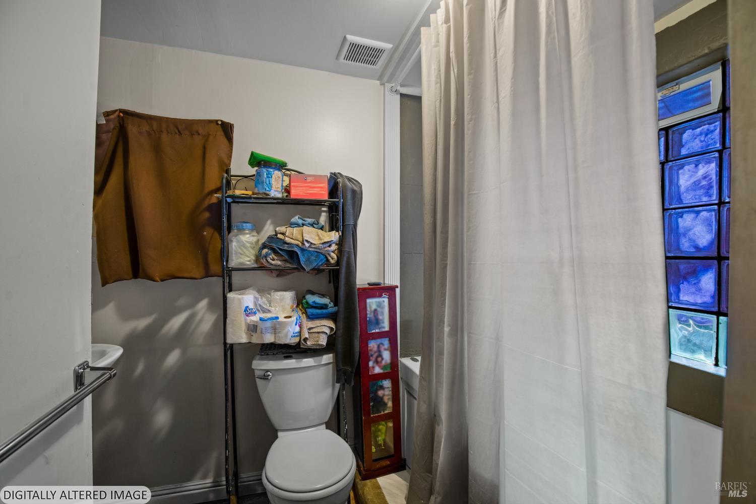 Bathroom with a toilet, storage rack, shower curtain, and blue glass-block window.