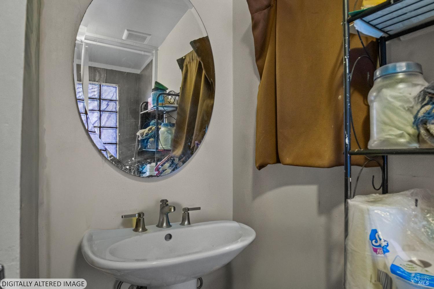 Compact bathroom sink beneath an oval mirror, with a brown curtain and storage shelves.