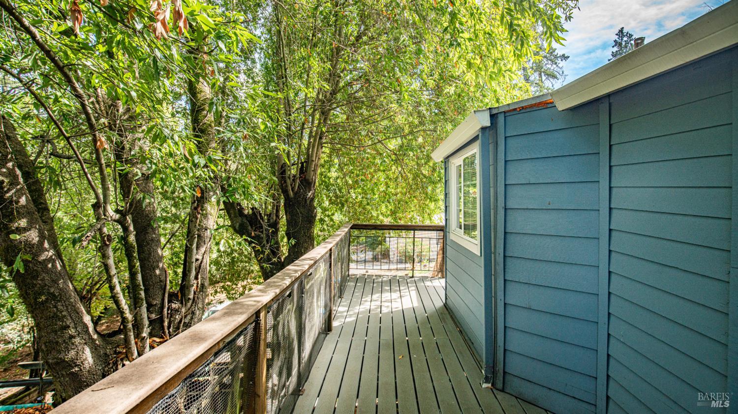 Elevated wooden deck beside a blue-sided home, surrounded by mature trees.