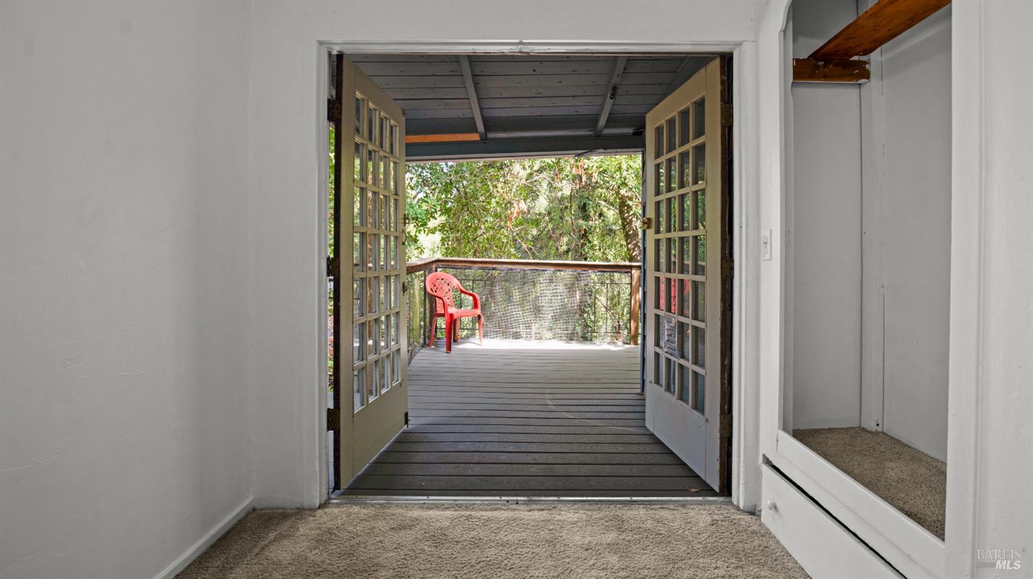 Empty carpeted room with open double doors leading to a covered deck overlooking trees.