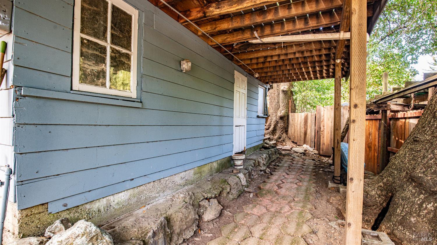 Covered side walkway beside a blue-sided house, with brick paving and large trees.