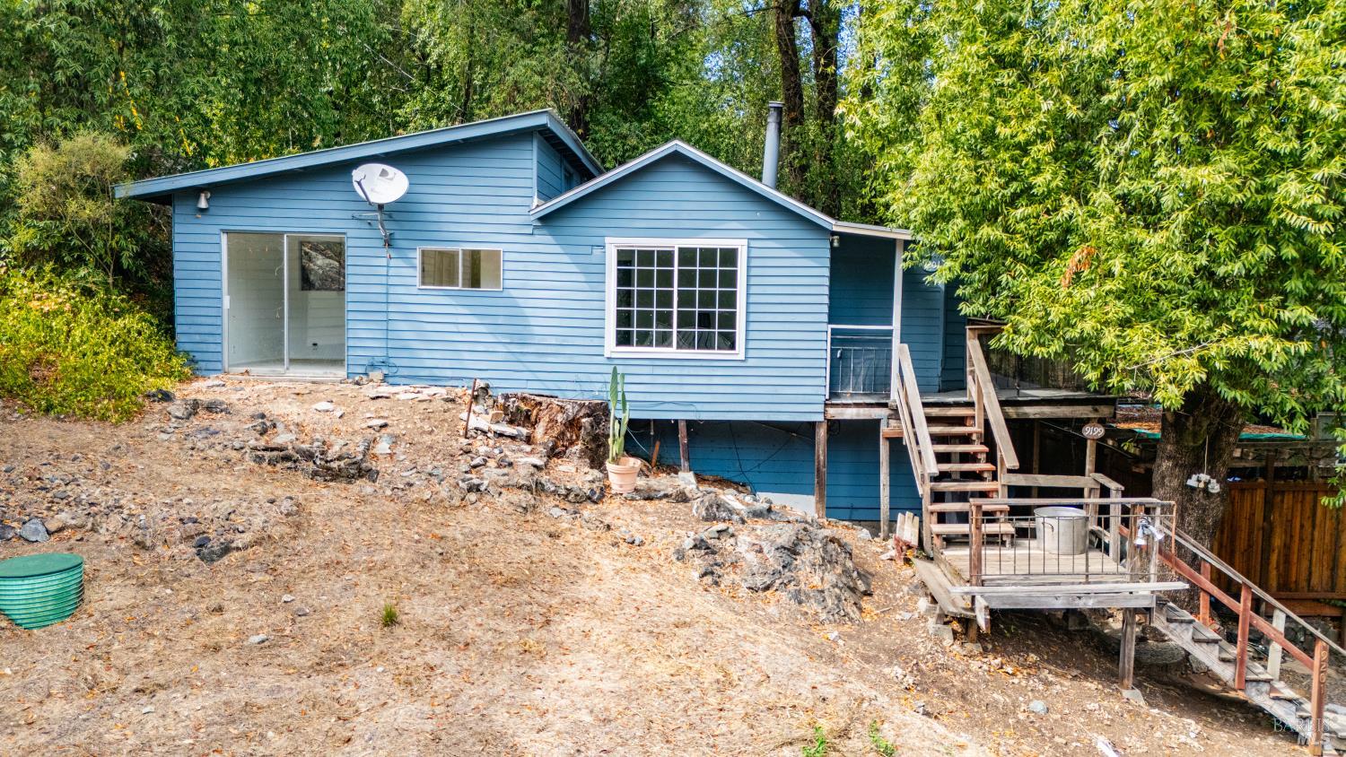 Blue house on a rocky slope, with a raised wooden deck and stairs among trees.