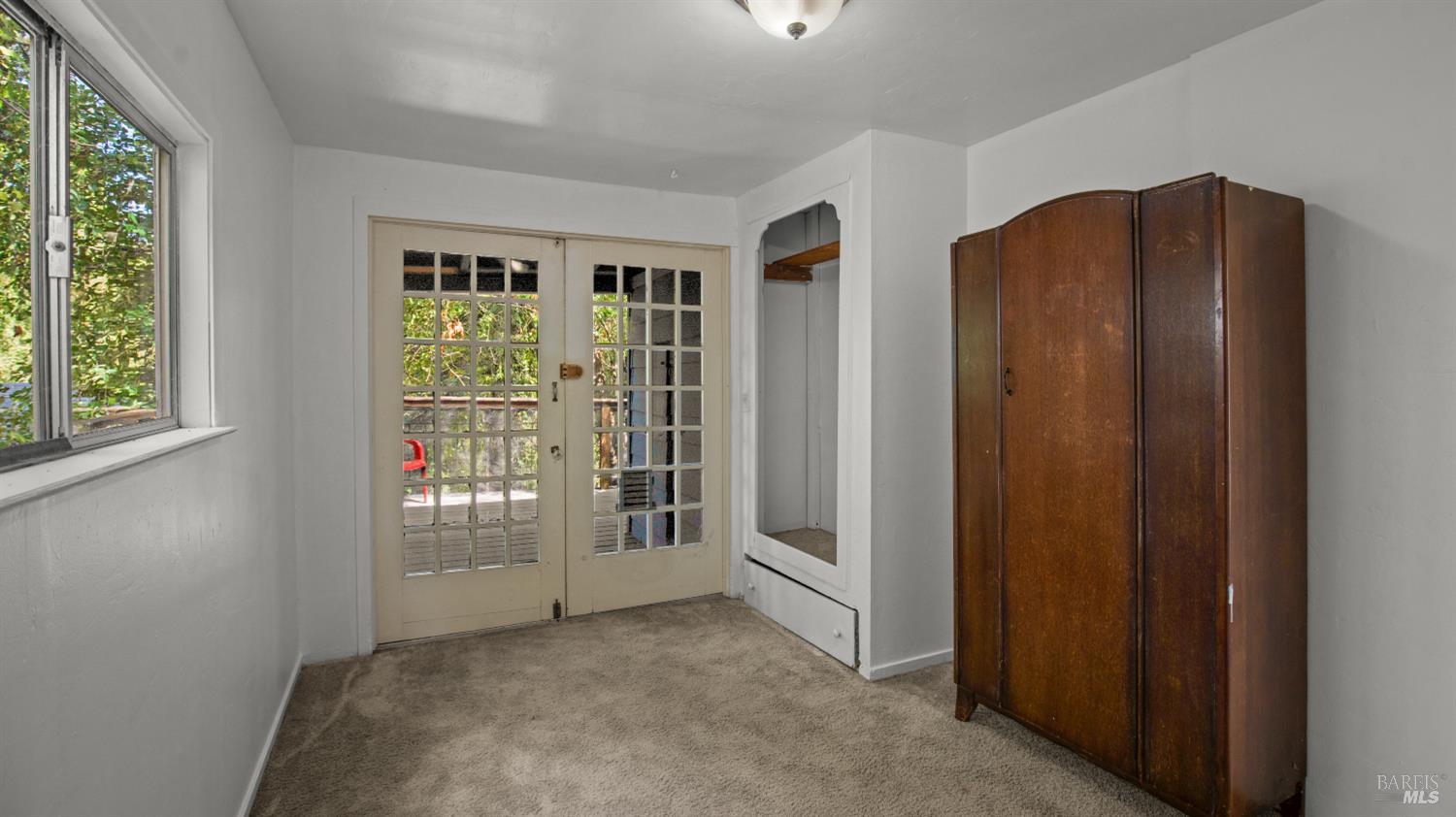 Carpeted bedroom with a wooden wardrobe, French doors to a deck, and a window overlooking greenery.