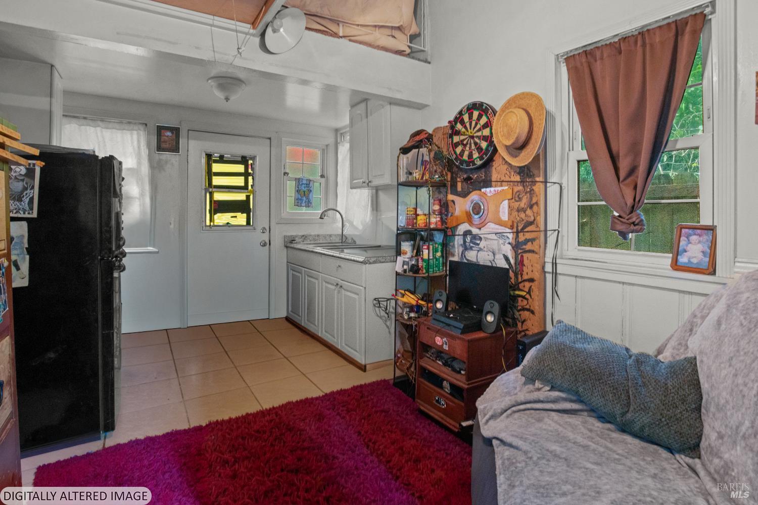 Furnished living room with a small kitchenette, dartboard, and window overlooking greenery.