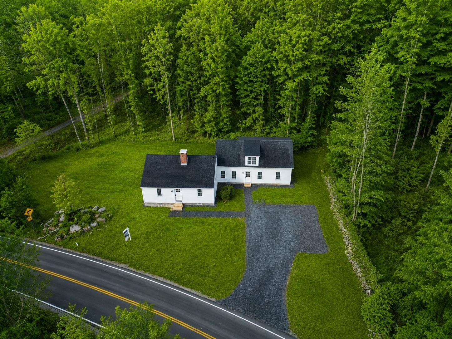 Aerial view of two white buildings and a gravel driveway on a wooded lot beside a road.
