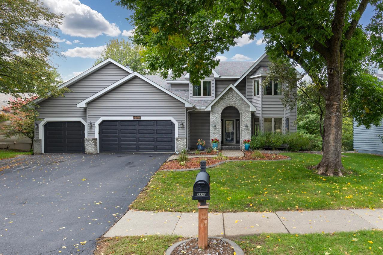 Gray two-story home with a three-car garage, stone entry, and mature trees.