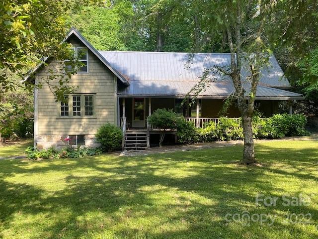 Gray-green house with a steep gable, metal roof, and covered porch among leafy trees.