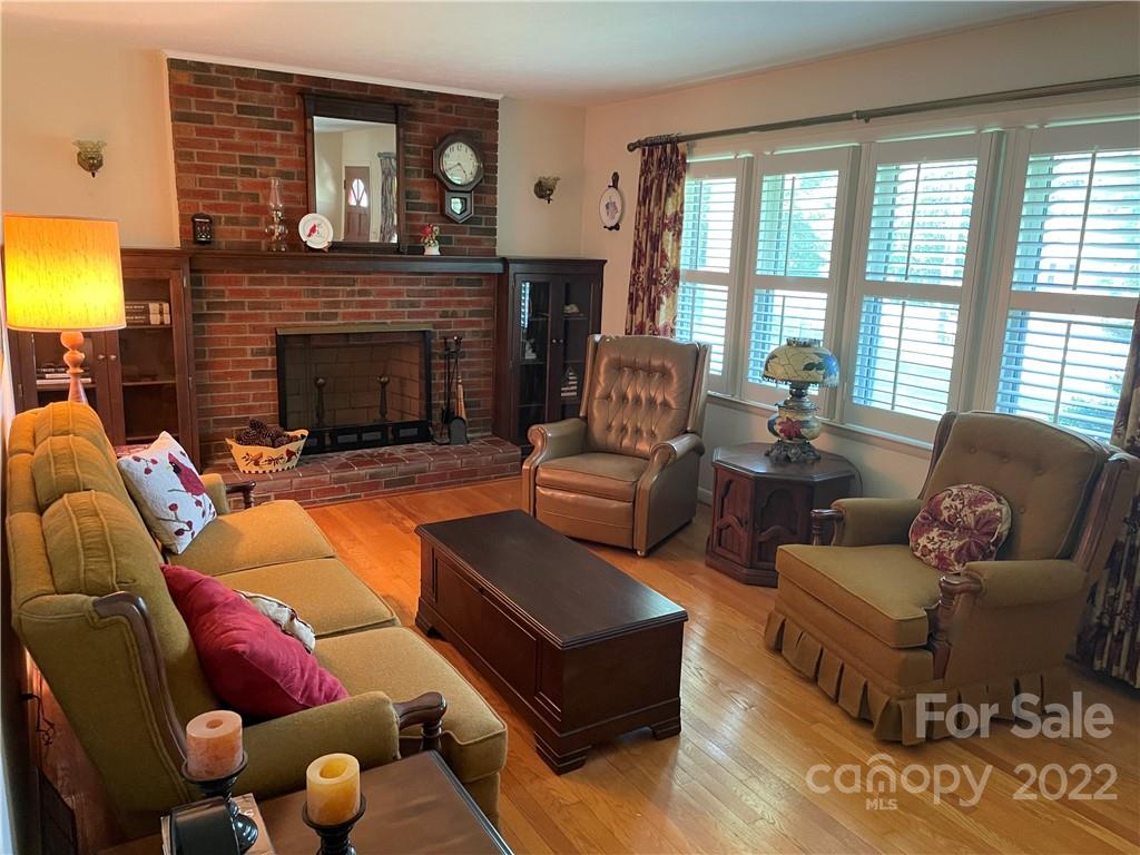 Furnished living room with a brick fireplace, wood floors, and a wall of shuttered windows.