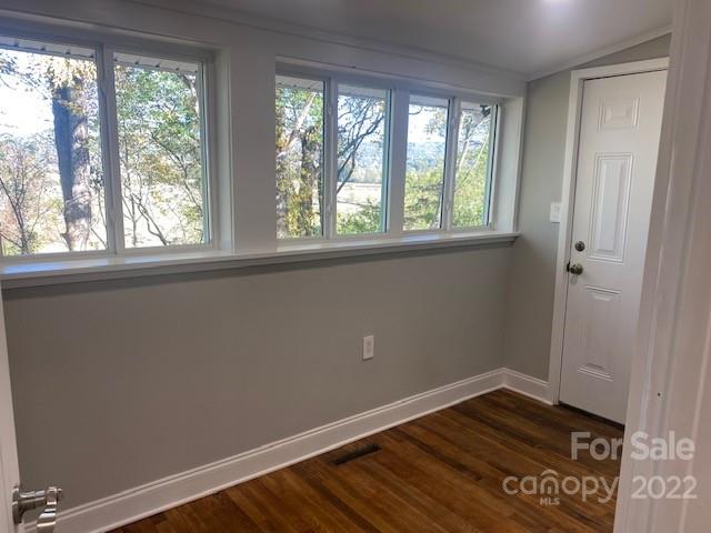 Vacant room with dark wood floors, gray walls, and wide windows overlooking trees.