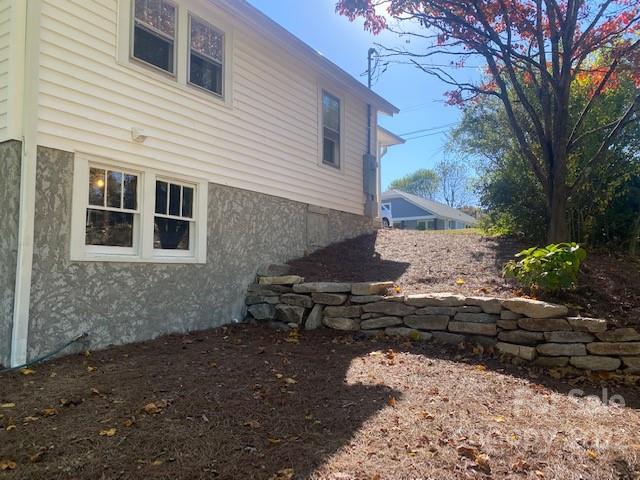 House side with pale siding and stone foundation beside a sloped yard and low retaining wall.