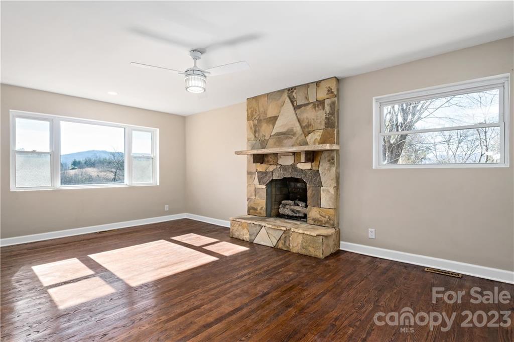 Vacant living room with a large stone fireplace, dark hardwood floors, and a wooded hill visible through the window.