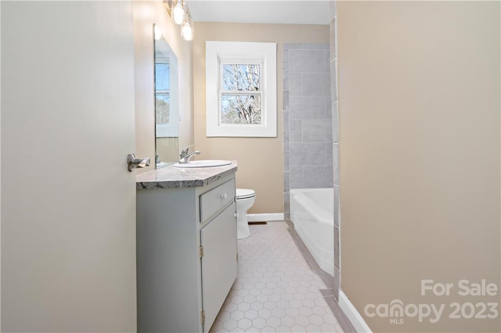 Bathroom with gray vanity, white bathtub, gray tiled wall, and a window overlooking trees.