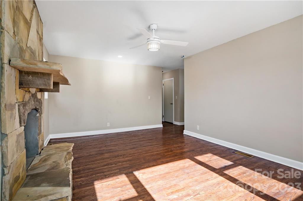 Empty living room with a stone fireplace, dark wood floors, and sunlight across the floor.