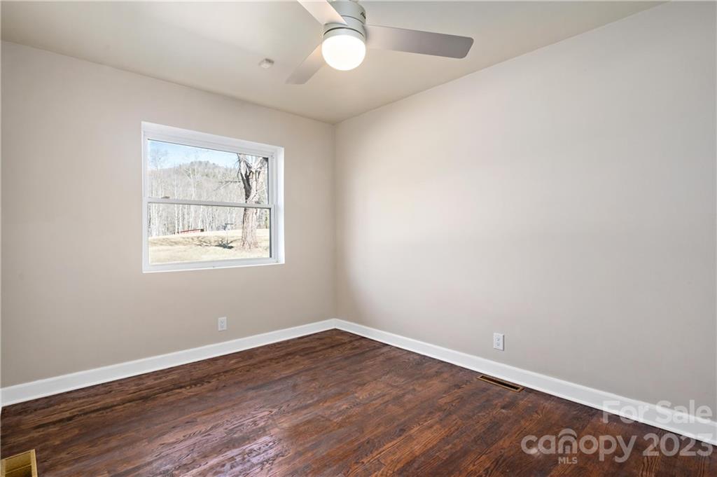 Empty bedroom with dark wood flooring, a window overlooking bare trees, and a ceiling fan.