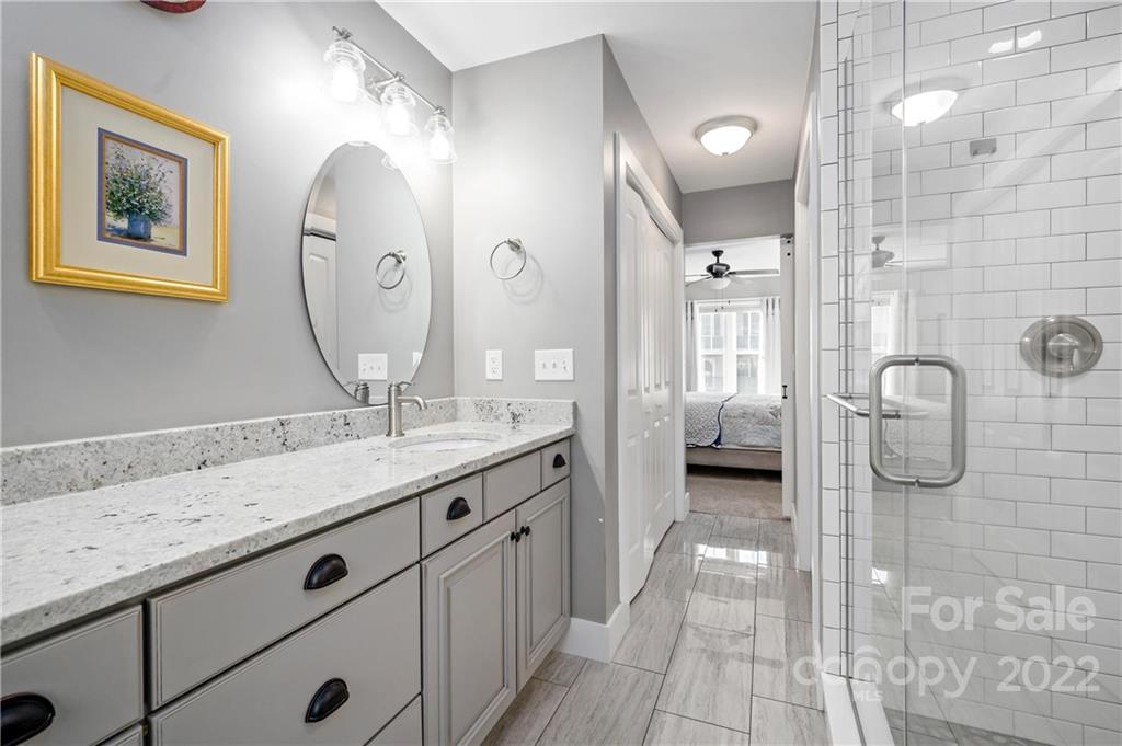 Bathroom with a long stone vanity and glass shower; a bedroom is visible through the doorway.