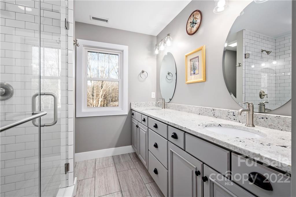 Bathroom with a gray double vanity, oval mirrors, and a glass-enclosed subway-tile shower.
