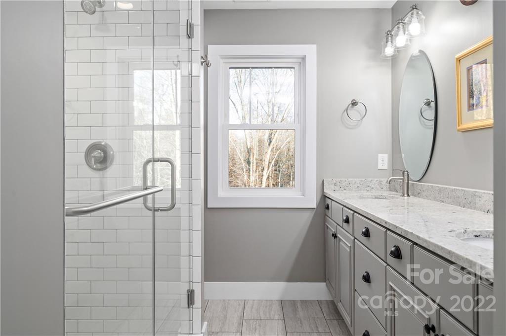 Bathroom with a glass subway-tile shower, gray vanity, stone countertop, and window overlooking trees.