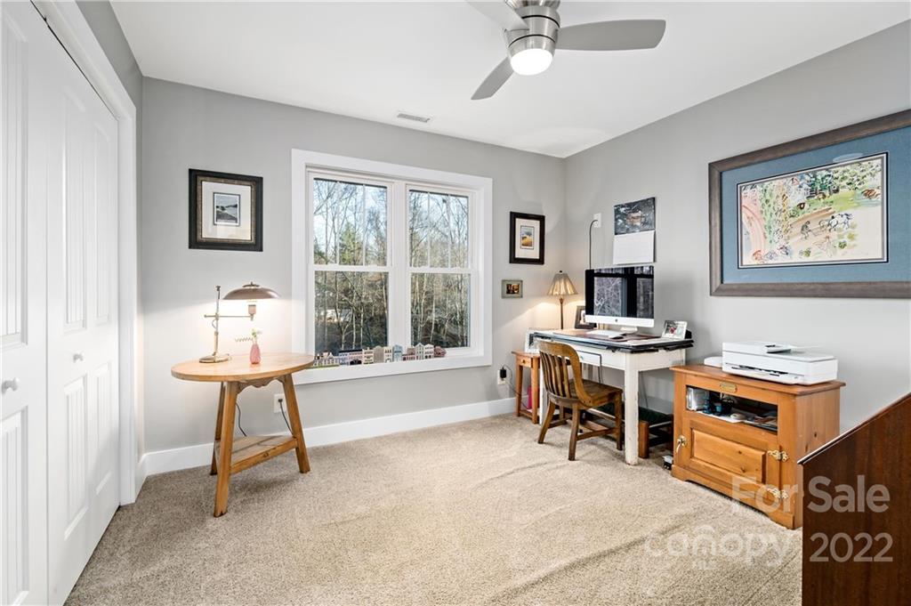 Home office with a large window overlooking leafless trees, a white desk, and beige carpet.