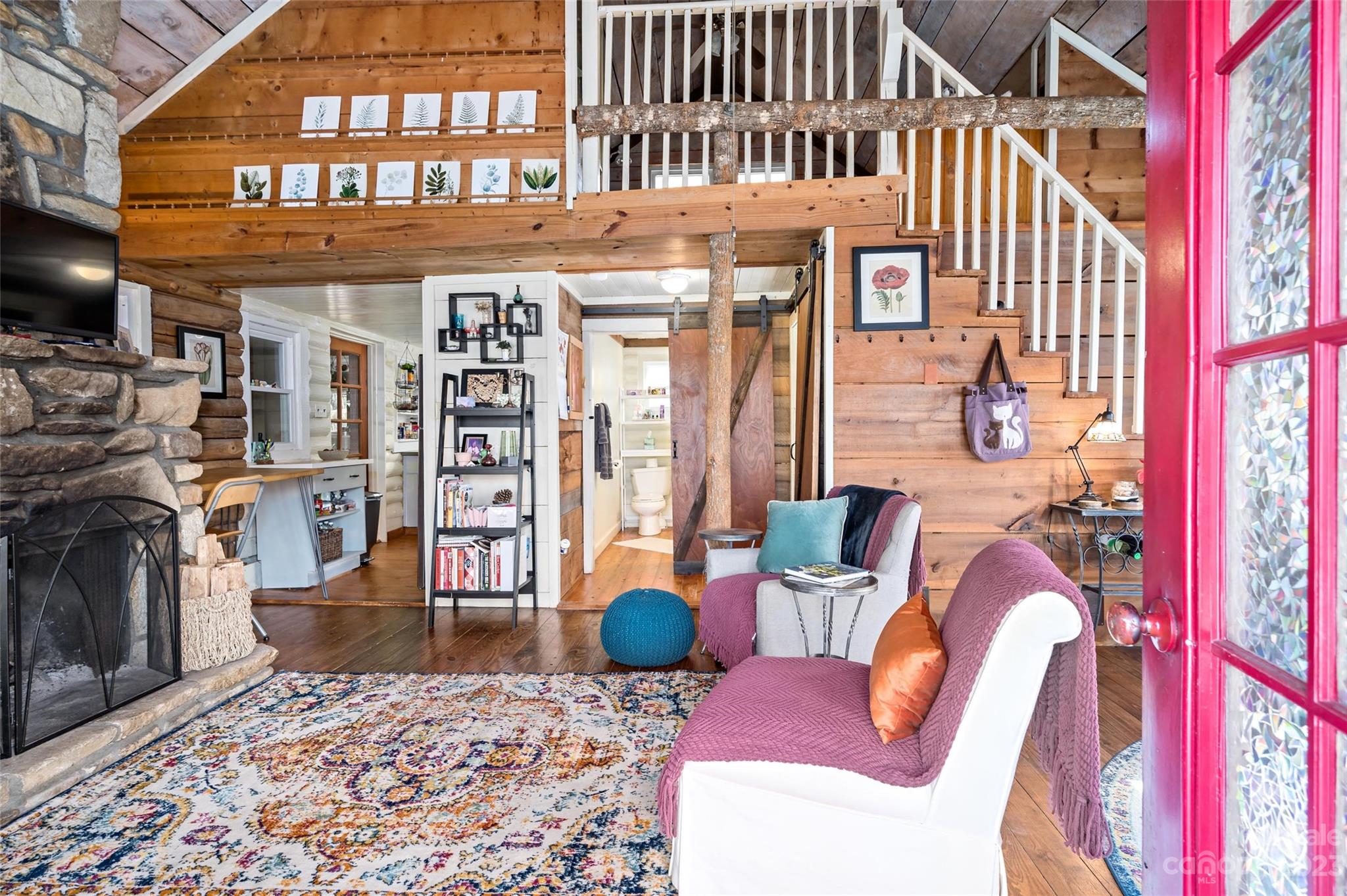 Cabin living room with a stone fireplace, loft, white-railed staircase, and open red stained-glass door
