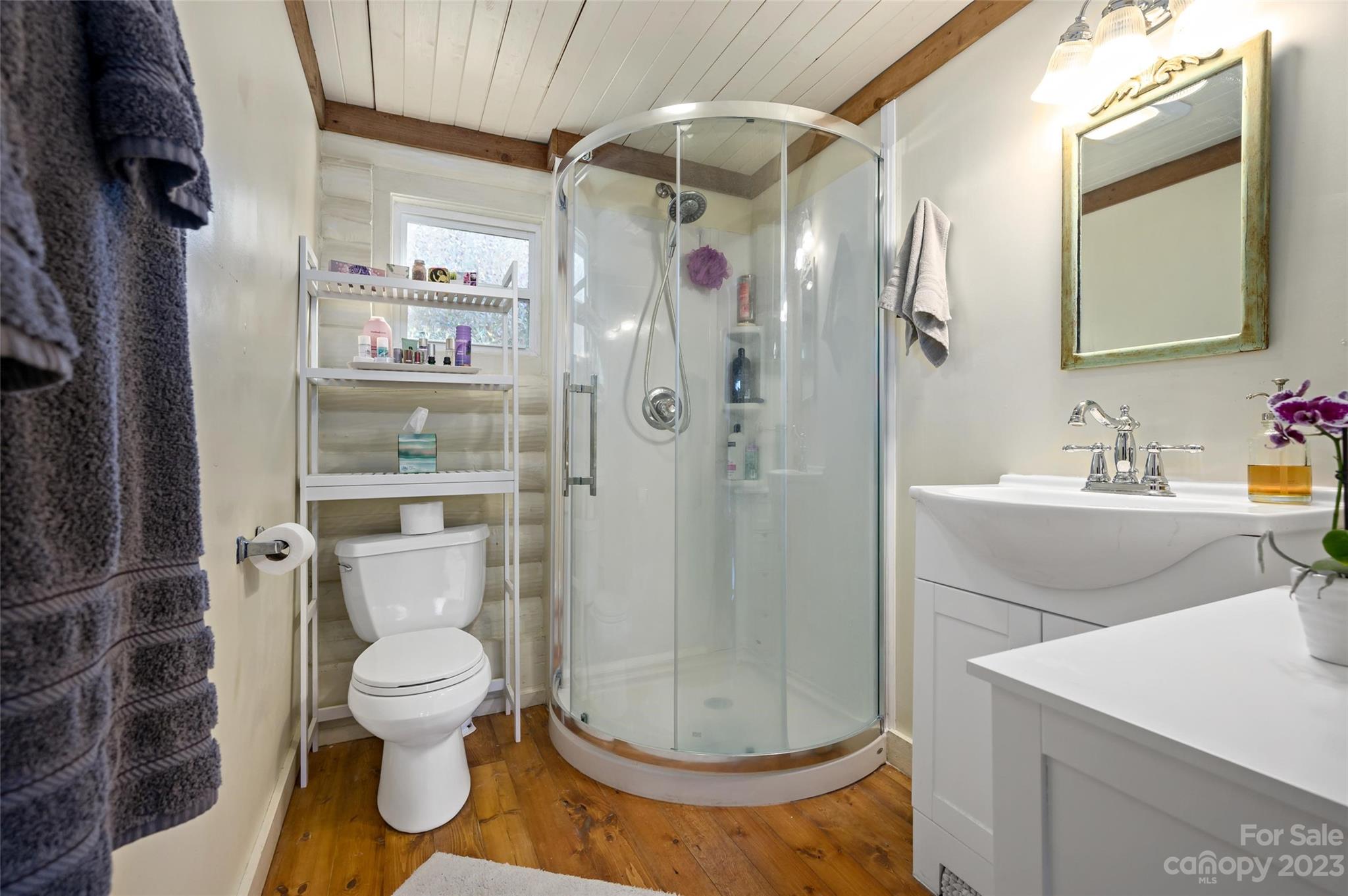 Bathroom with a curved glass shower, white vanity, wood floor, and exposed ceiling beams.