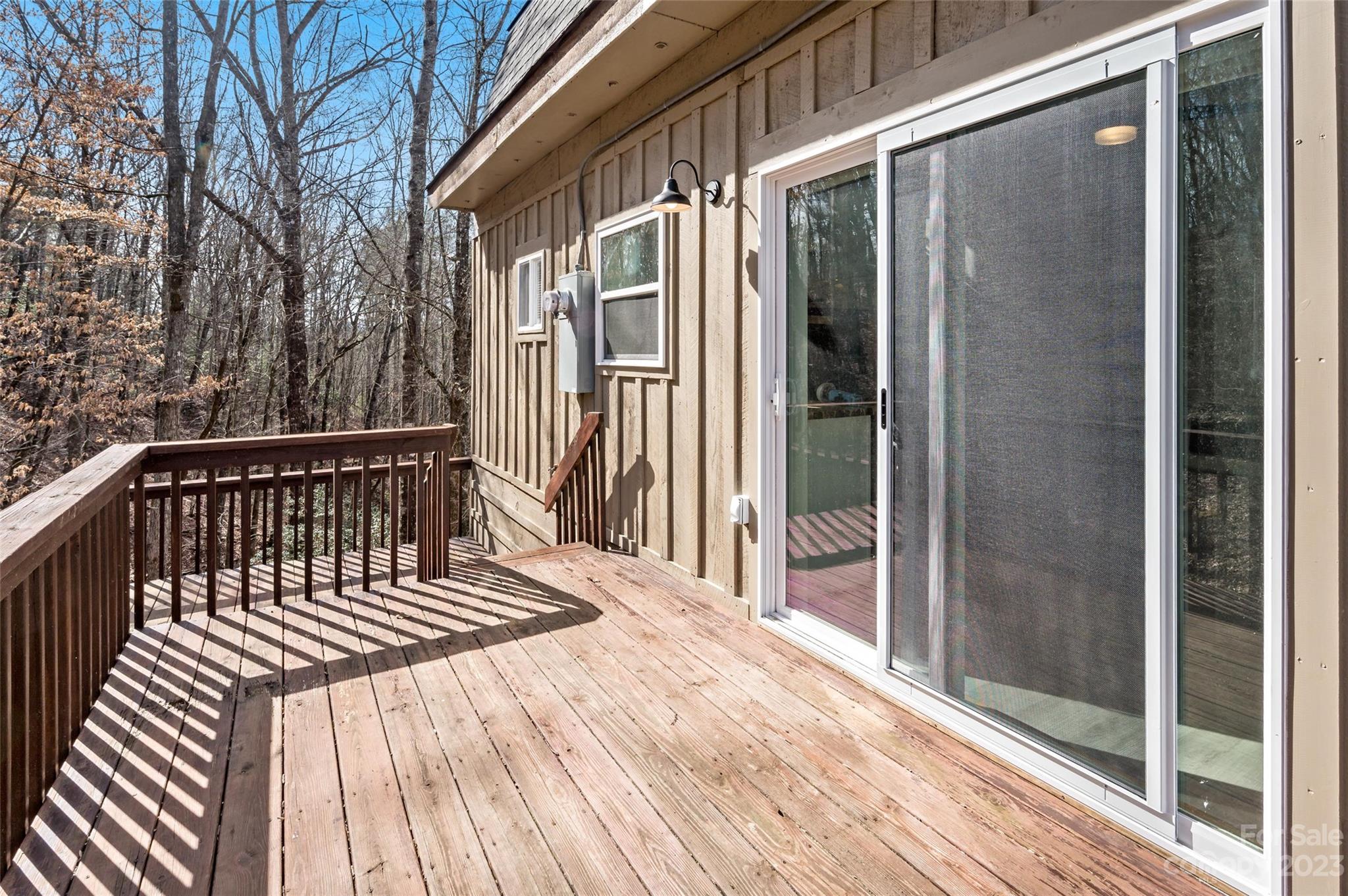Wooden deck beside a tan-sided home, overlooking leafless trees and accessed through a screened sliding door.