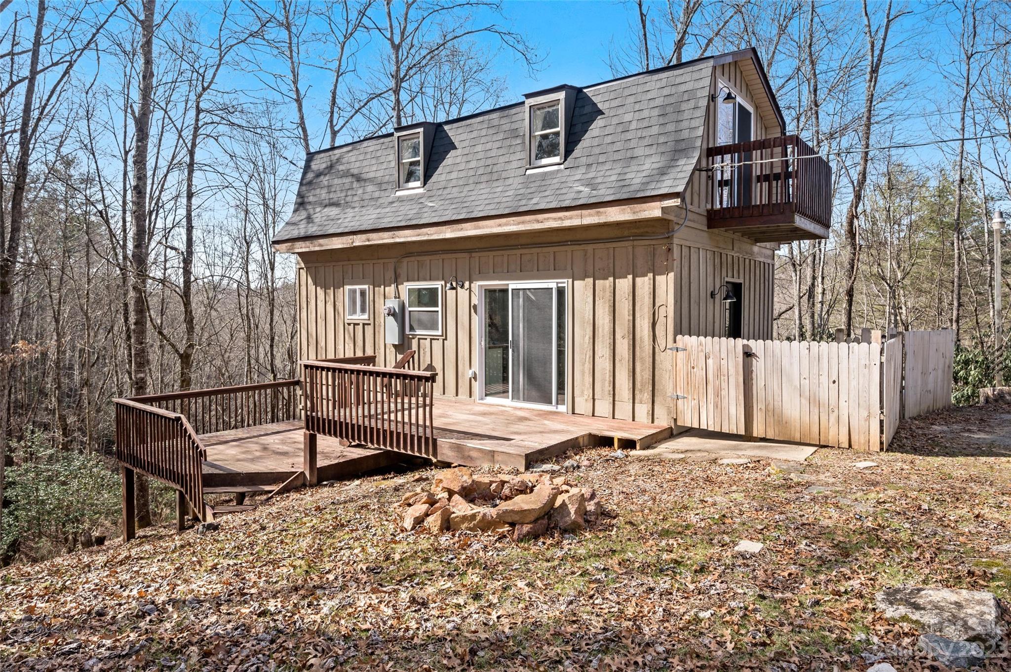Tan house with steep shingle roof, upper balcony, and wooden deck among bare trees.