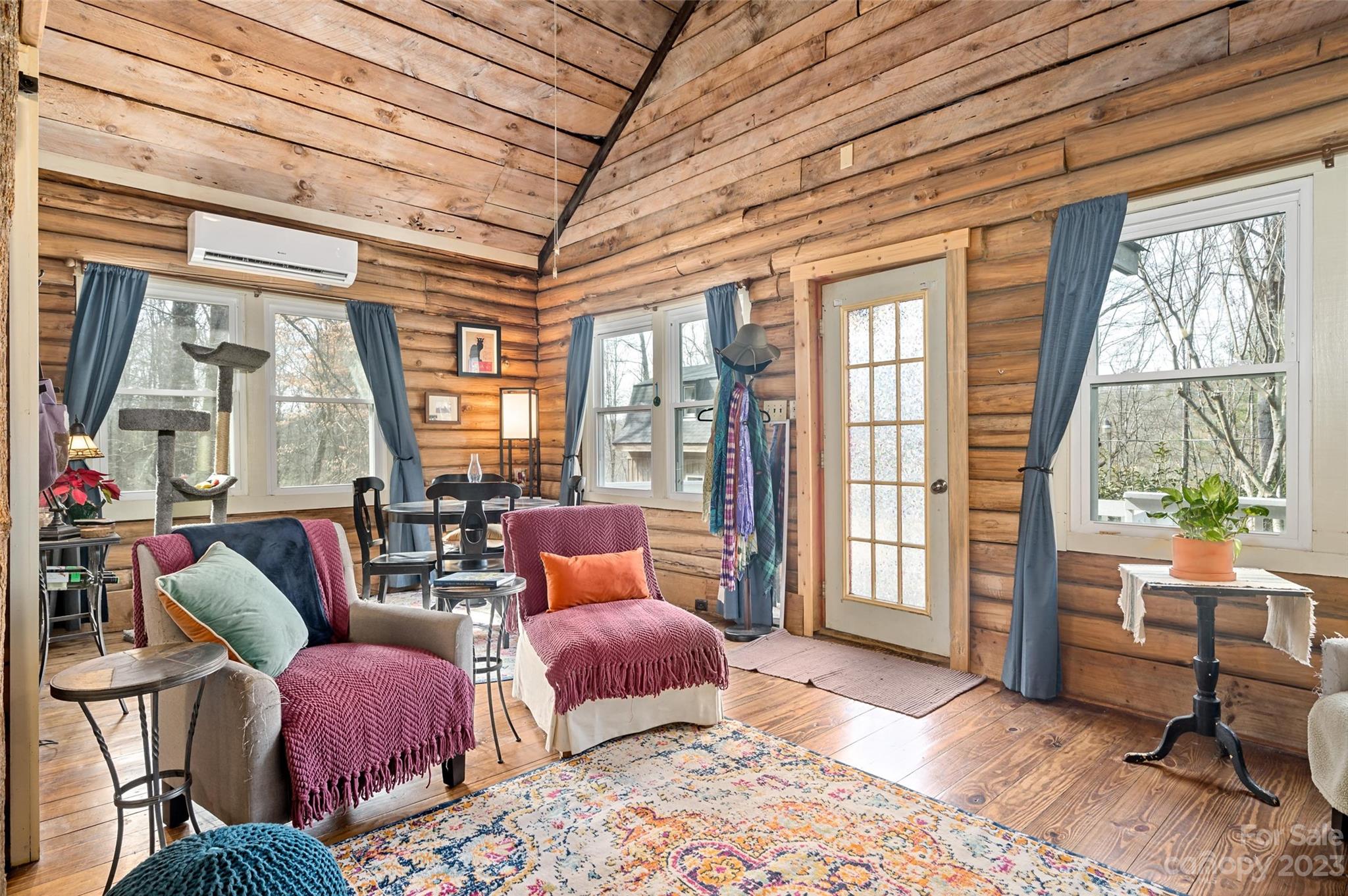 Furnished cabin living room with vaulted wood-plank ceiling, colorful rugs, and windows overlooking trees.