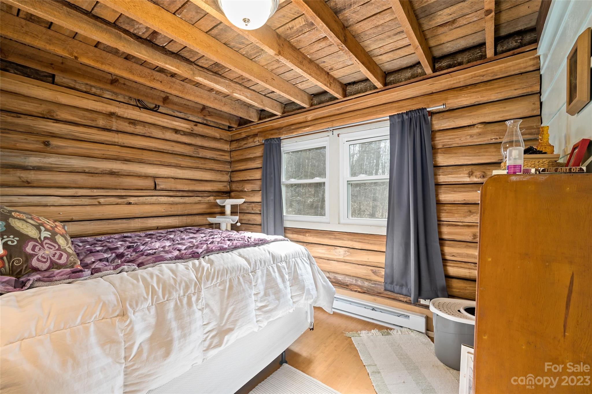 Log cabin bedroom with exposed timber walls and beams, a bed, and a double window with gray curtains.