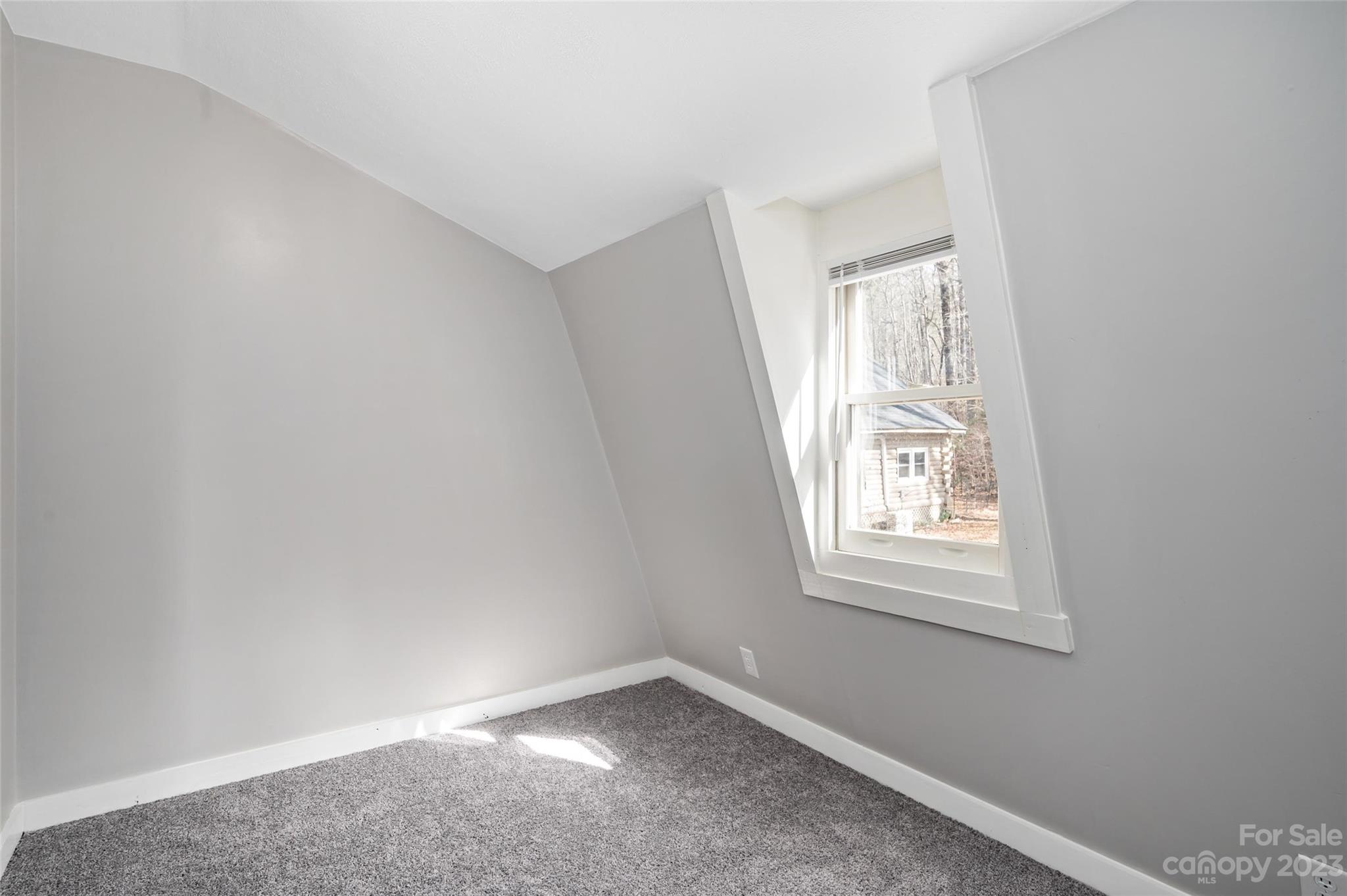 Empty carpeted room with sloped ceilings and a large window overlooking trees.