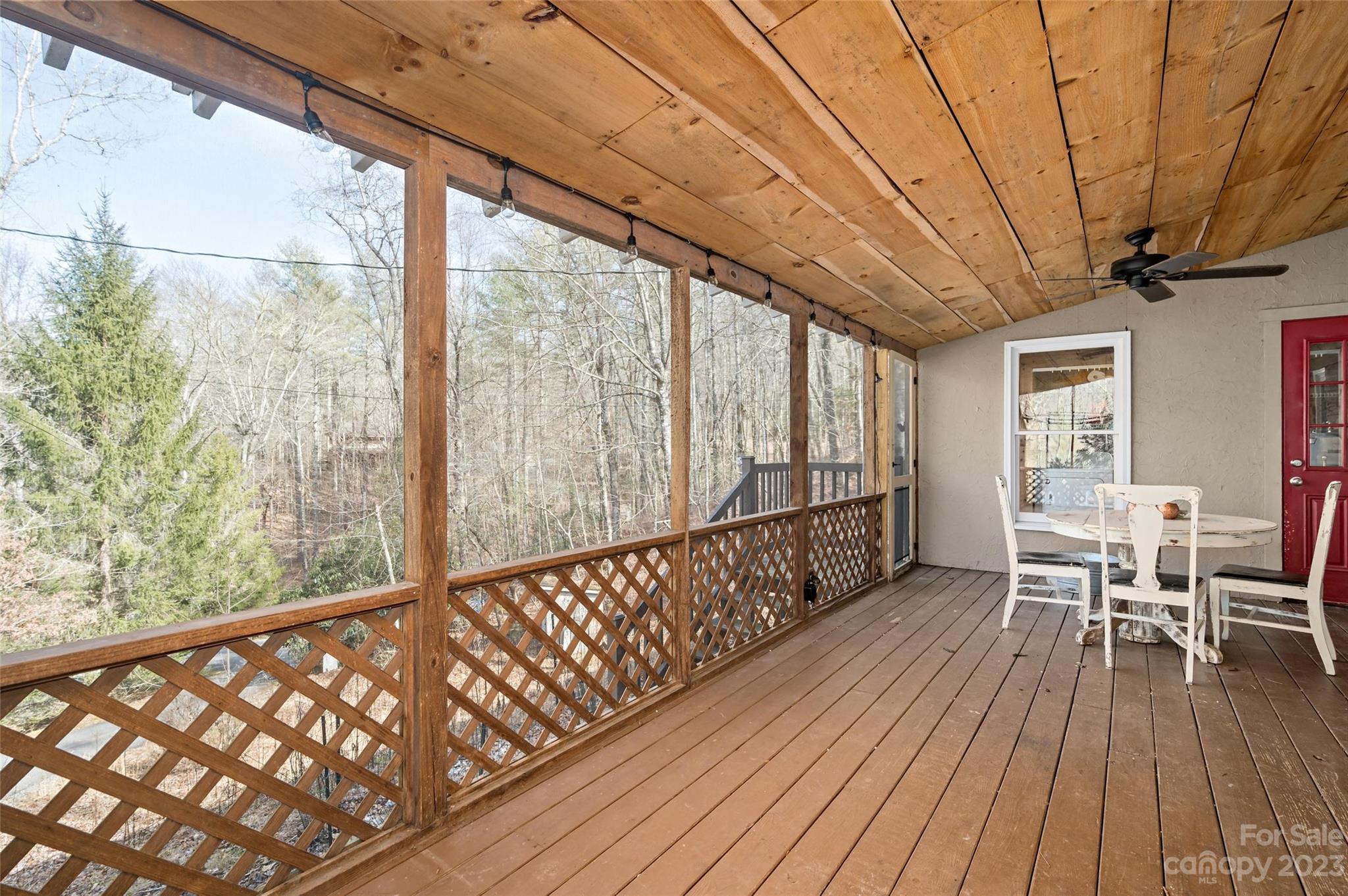 Covered wooden porch with a white dining set, lattice railing, and a view of leafless woods.