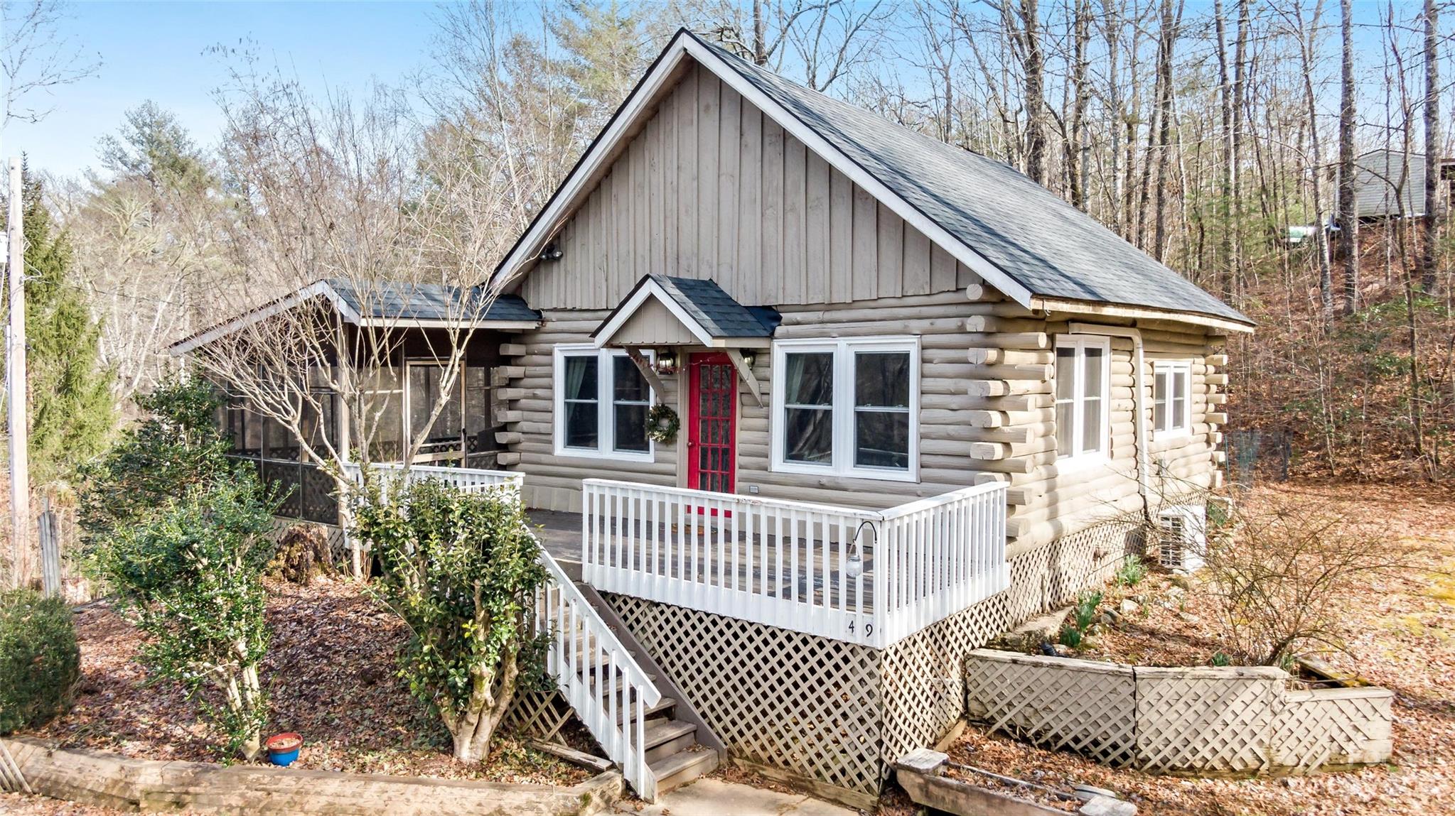 Log cabin with a red front door, white-railed porch, and screened porch among leafless trees.