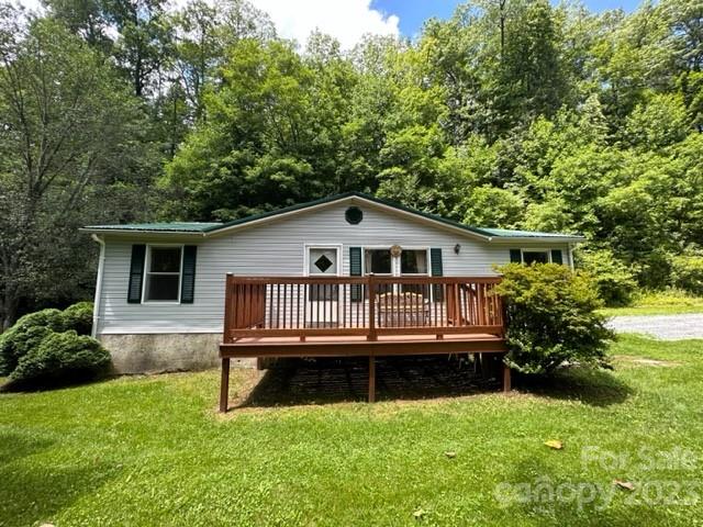 White single-story home with a raised wooden deck, grassy lawn, and dense trees behind it.