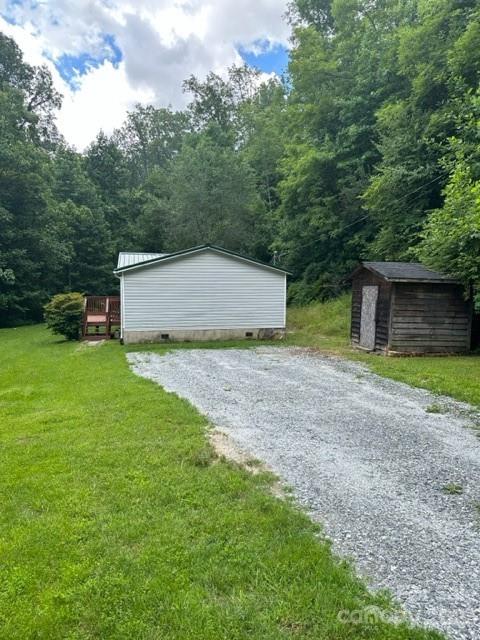 White manufactured home beside a gravel driveway, with a wooden shed and wooded surroundings.