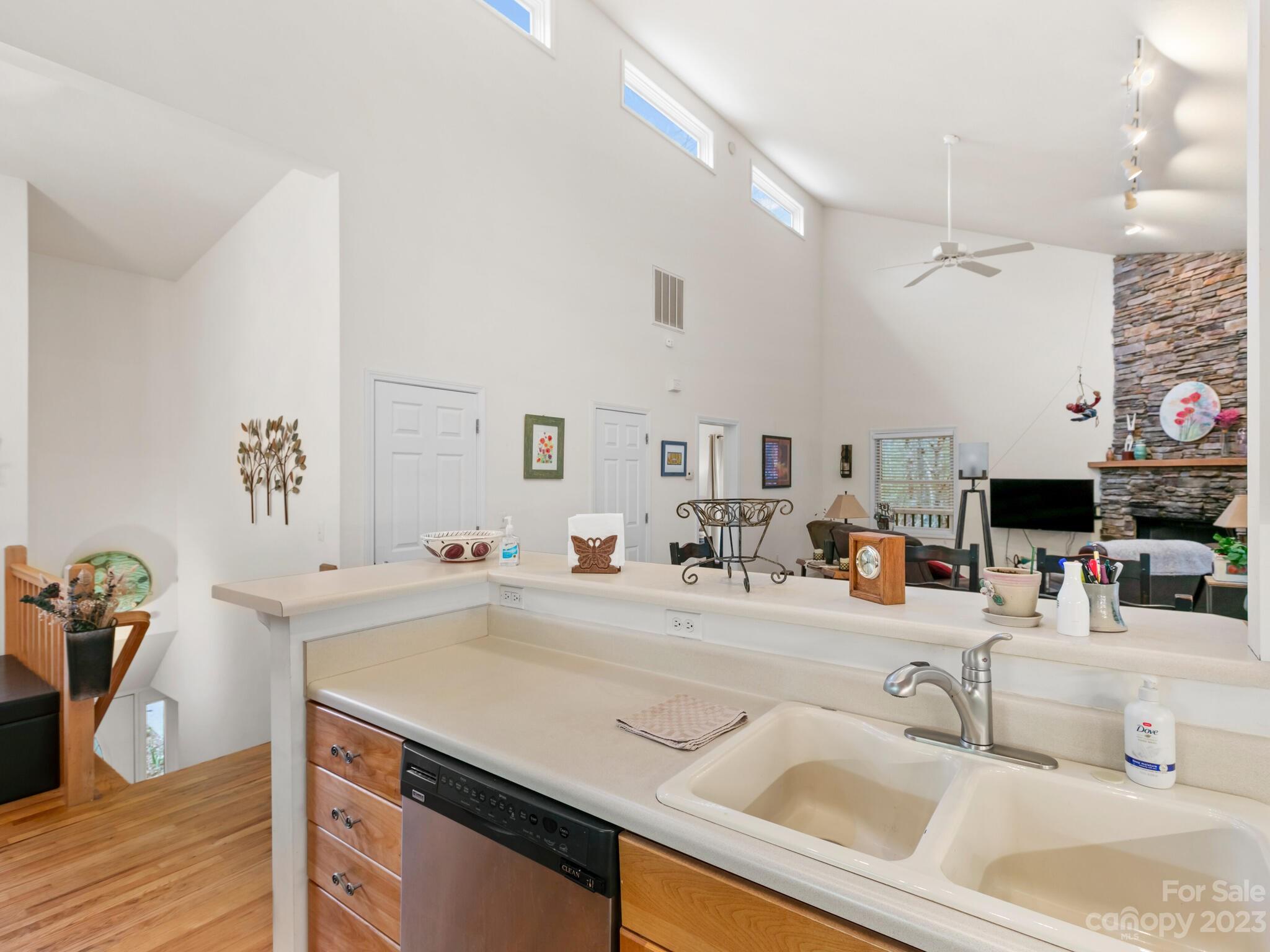 Kitchen counter with double sink and dishwasher overlooking a vaulted living room with a stone fireplace.