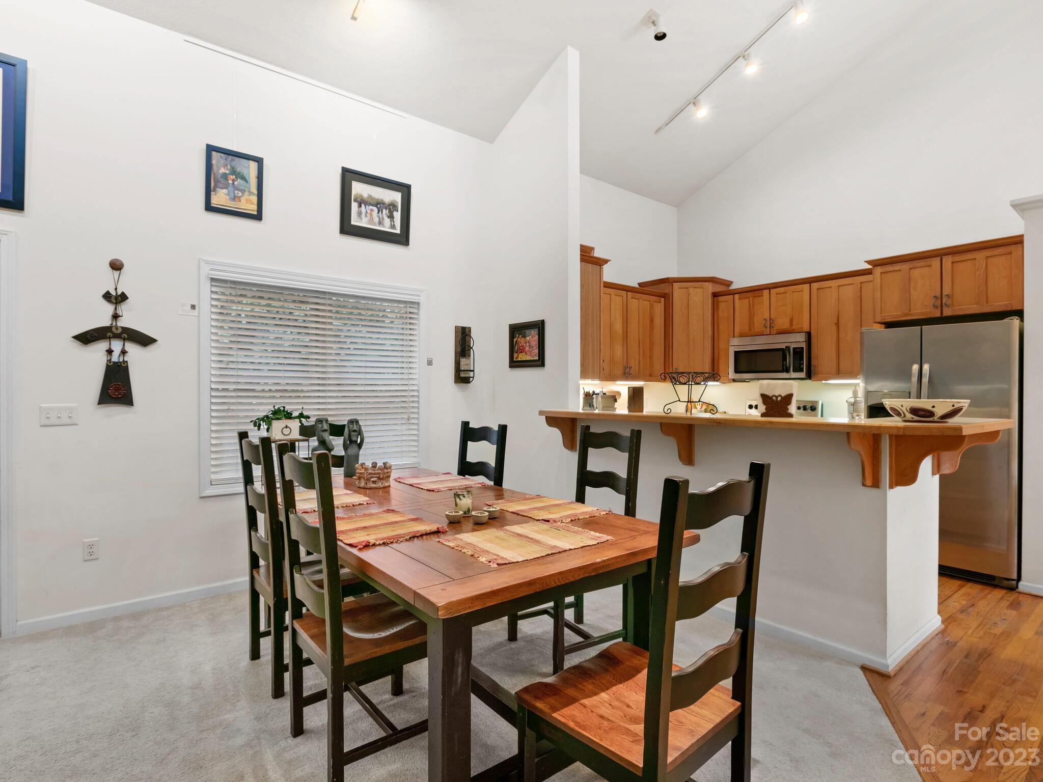 Dining area with a wooden table and black chairs beside a kitchen with honey-toned cabinets and stainless steel appliances.