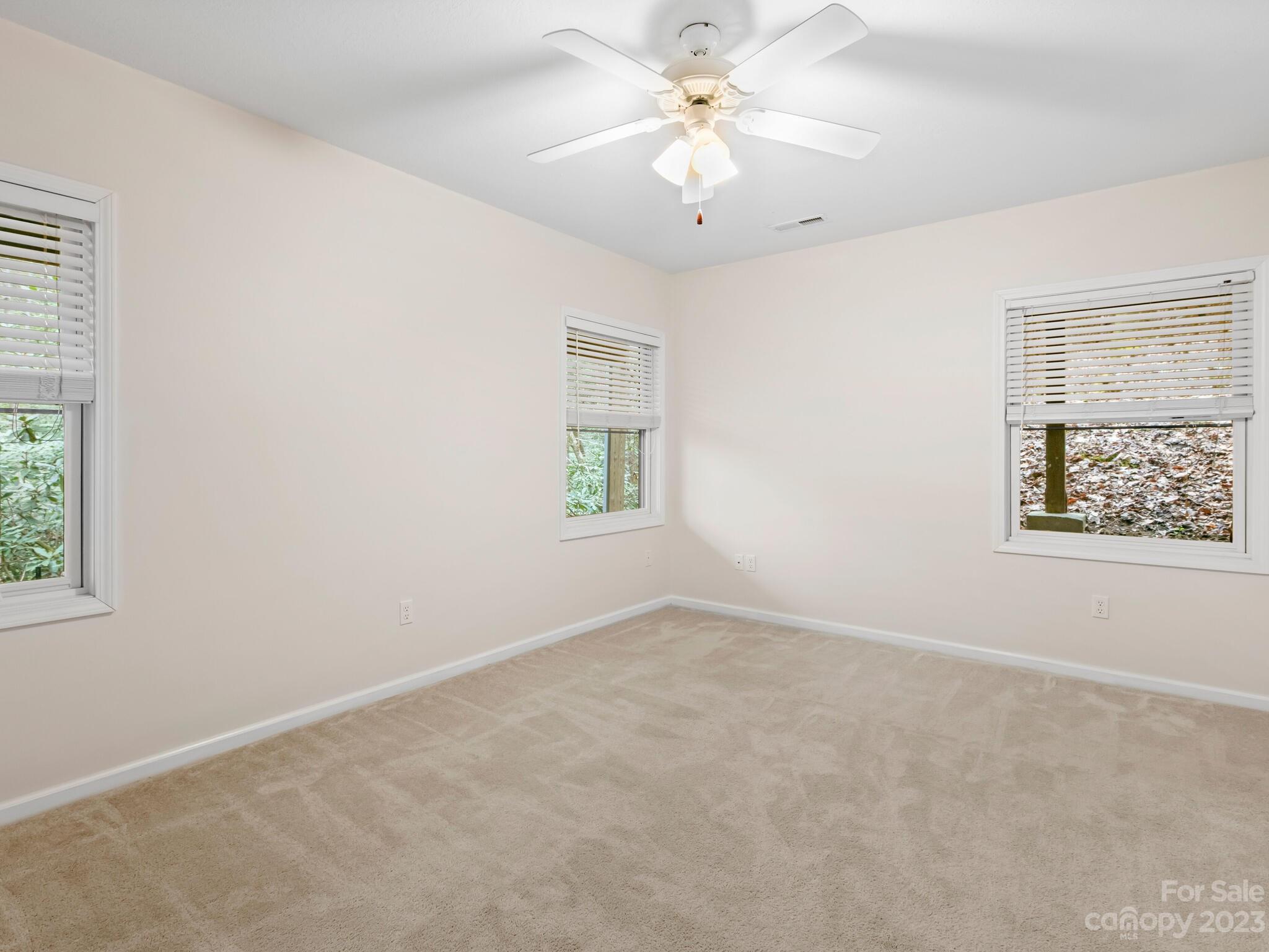 Empty carpeted bedroom with three blinded windows and a ceiling fan with lights.