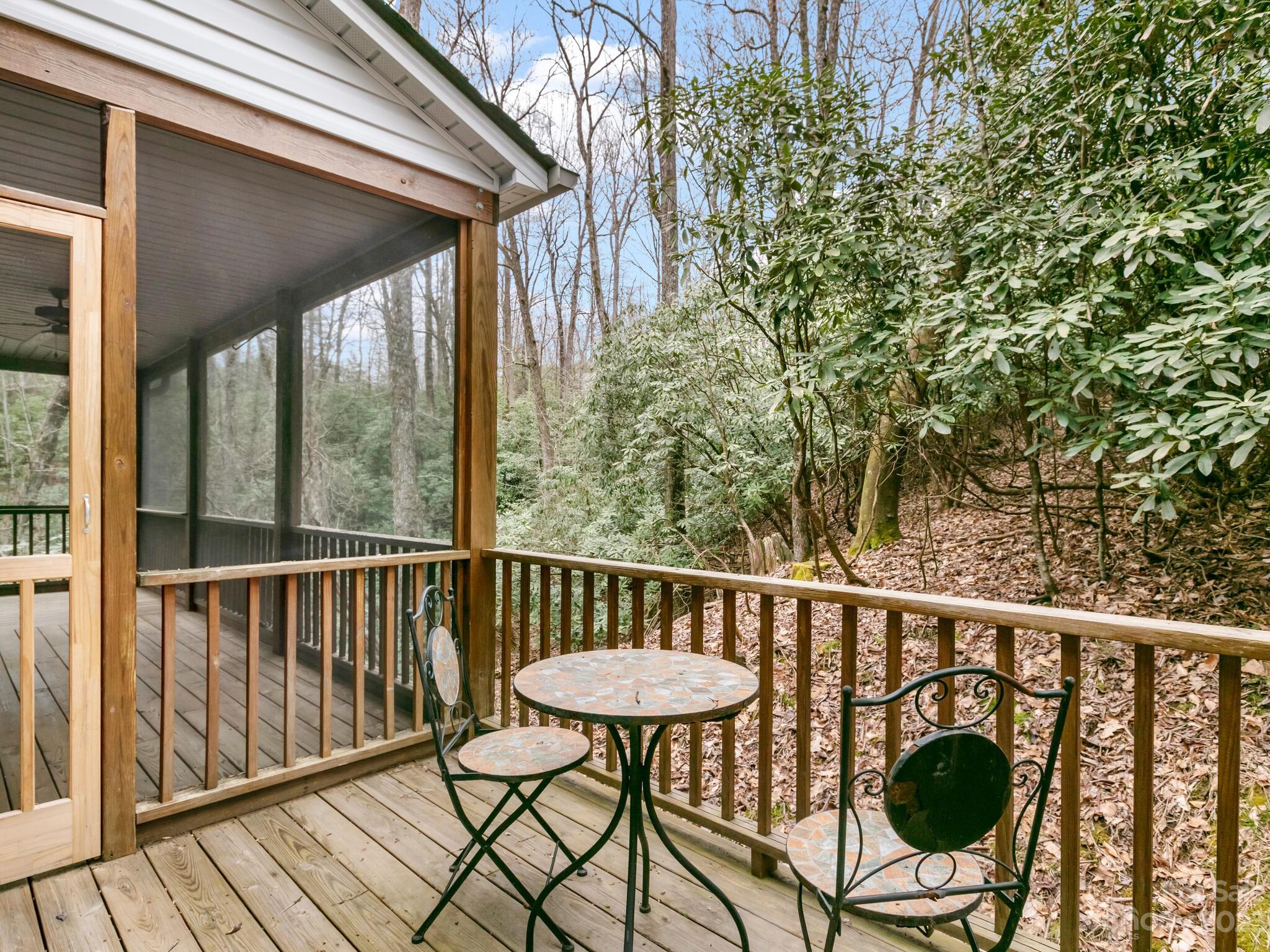 Wooden deck with mosaic patio table and chairs beside a screened porch, overlooking a wooded slope.