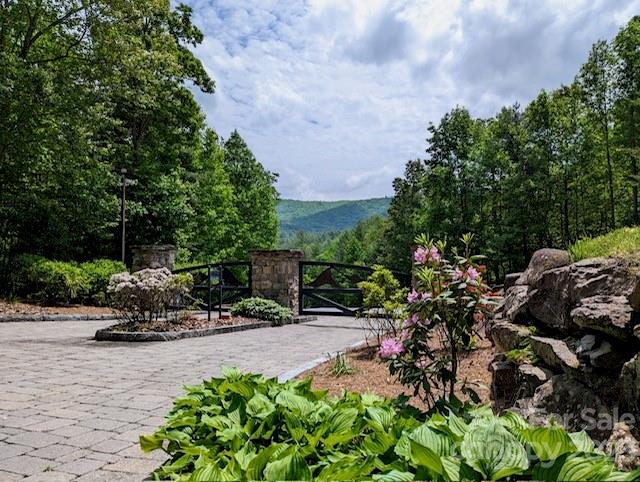Paved entrance with black gates and stone pillars, landscaped beds, and mountains in the distance.
