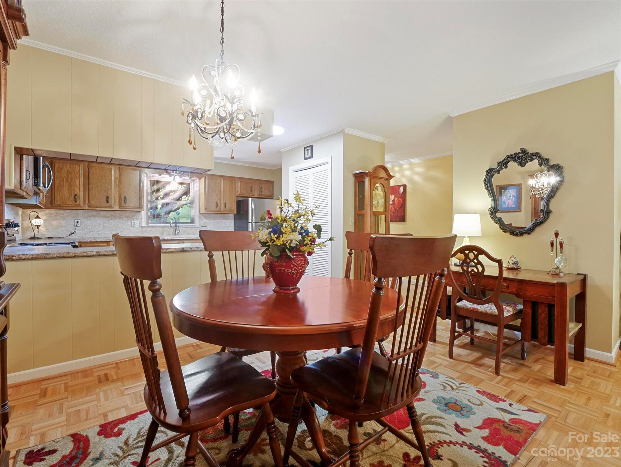 Dining area with a round wooden table, chandelier, patterned rug, and adjoining wood-cabinet kitchen.