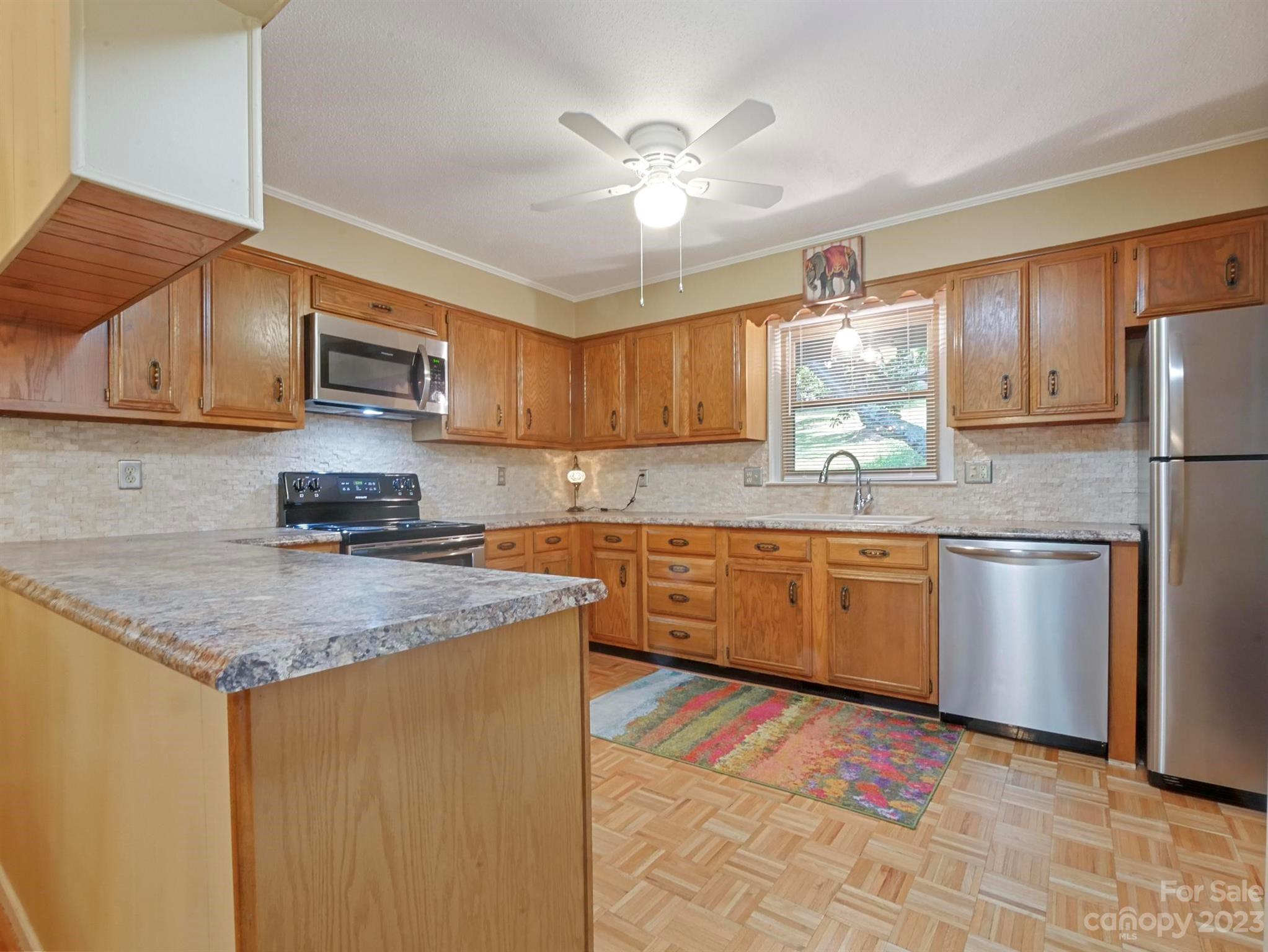 Kitchen with honey-toned wood cabinetry, stone counters, stainless steel appliances, and a colorful floor rug.