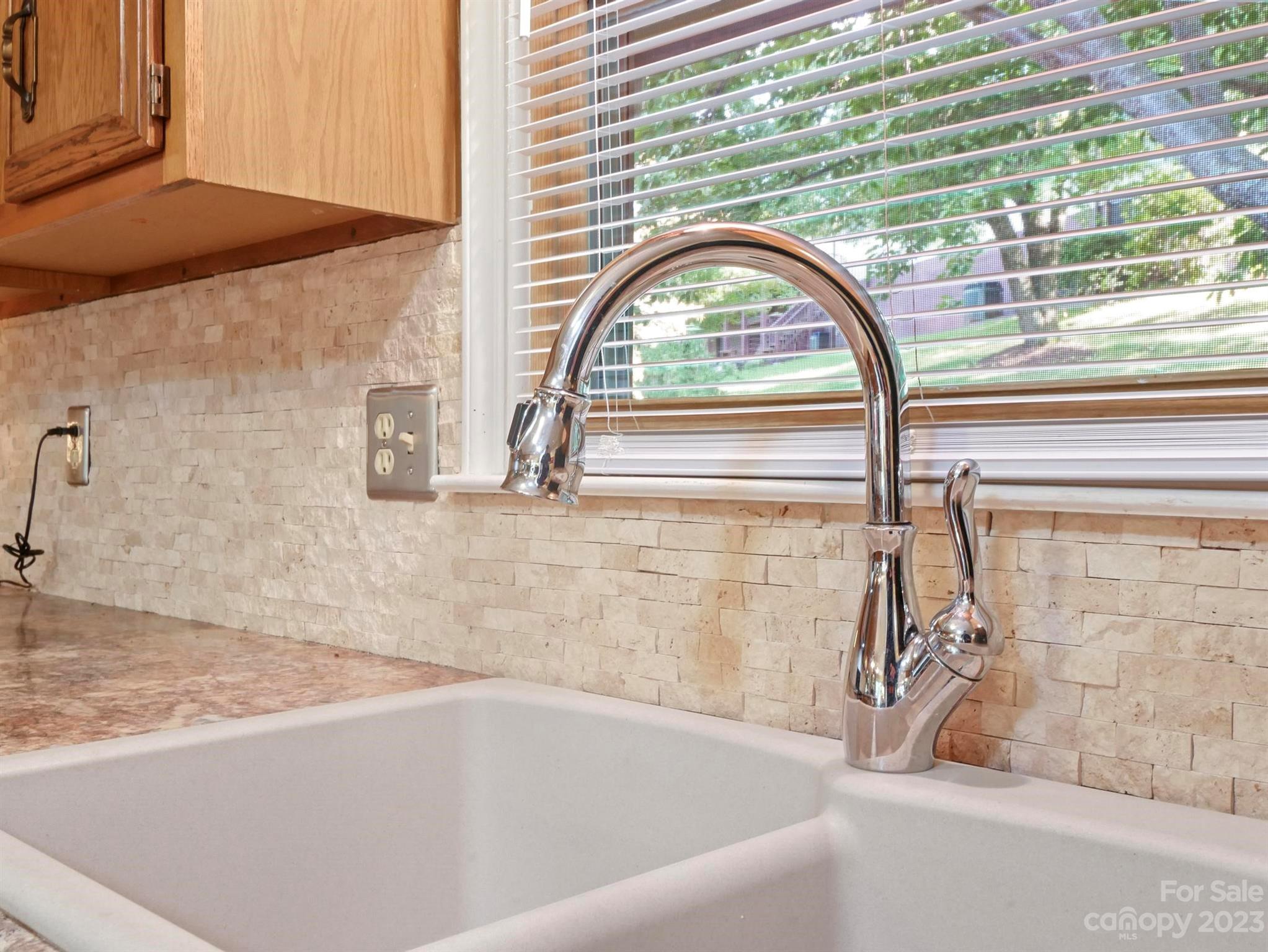 Kitchen sink with a tall chrome faucet, beige stone backsplash, and window blinds.