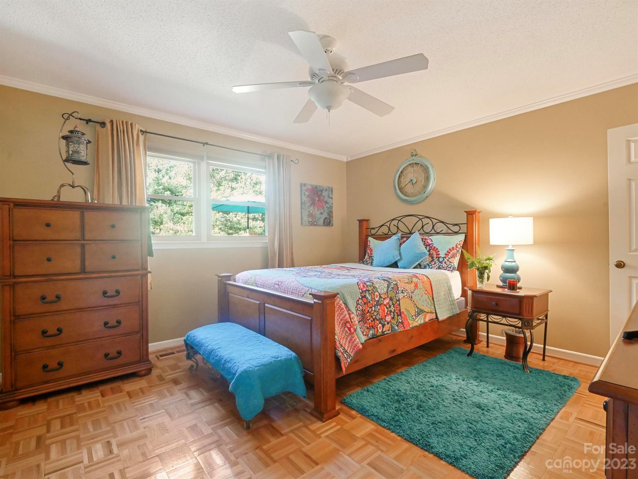 Furnished bedroom with colorful floral bedding, a teal rug, and daylight through two windows.