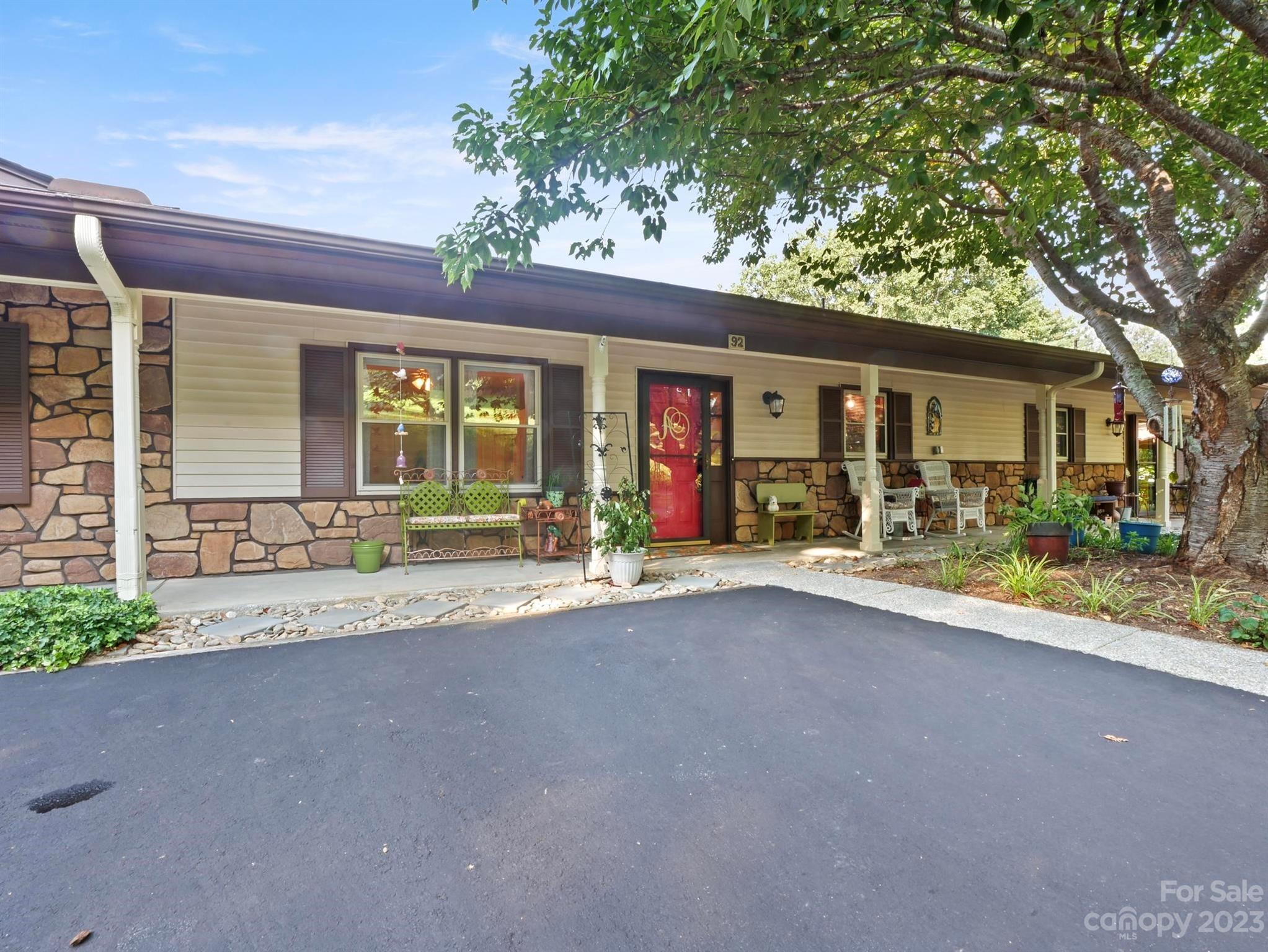 Single-story home with stone veneer, a red front door, and a covered porch beneath a large tree.