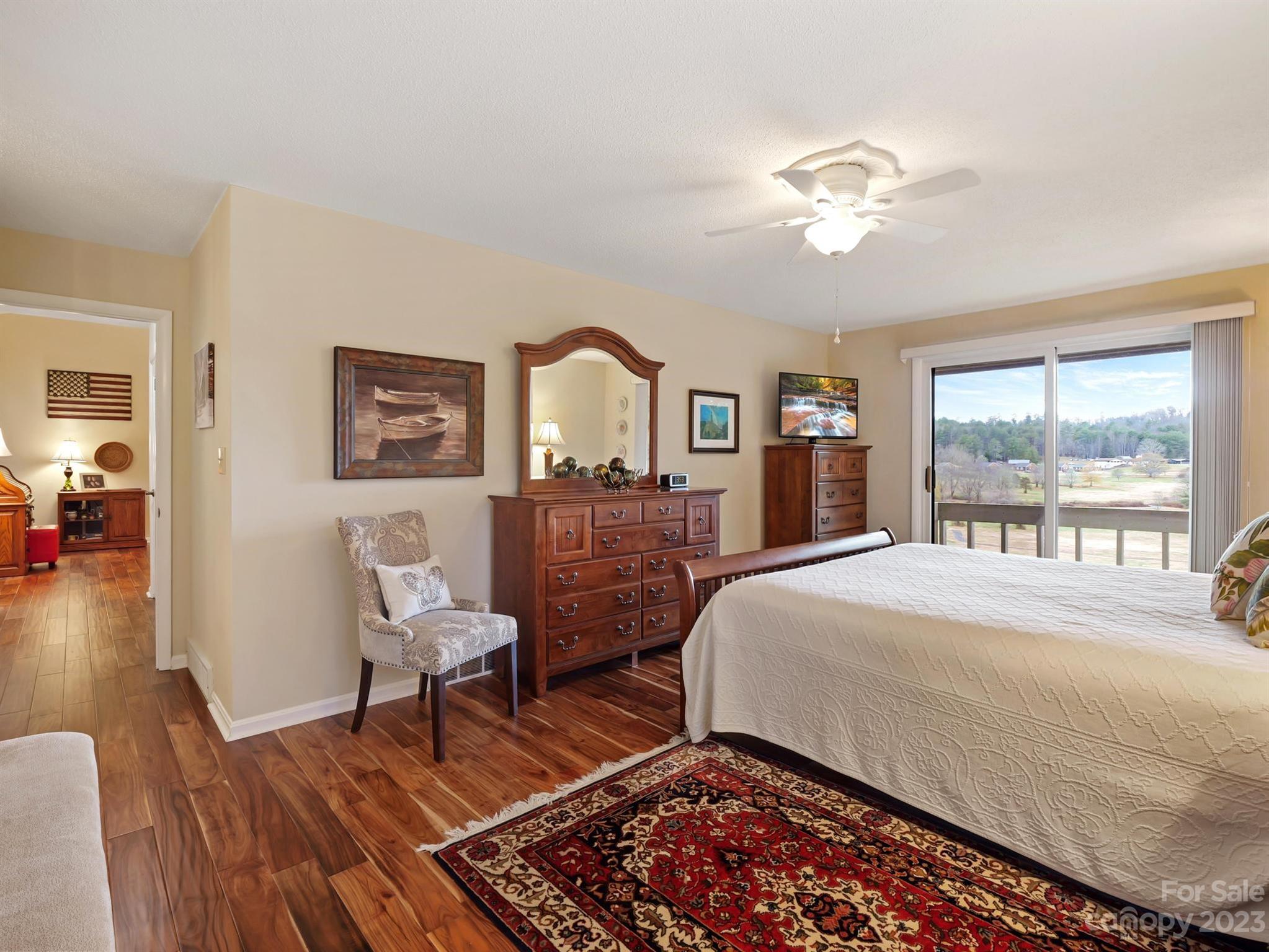 Furnished bedroom with a white quilted bed, dark wood furniture, and sliding doors to a balcony overlooking trees.