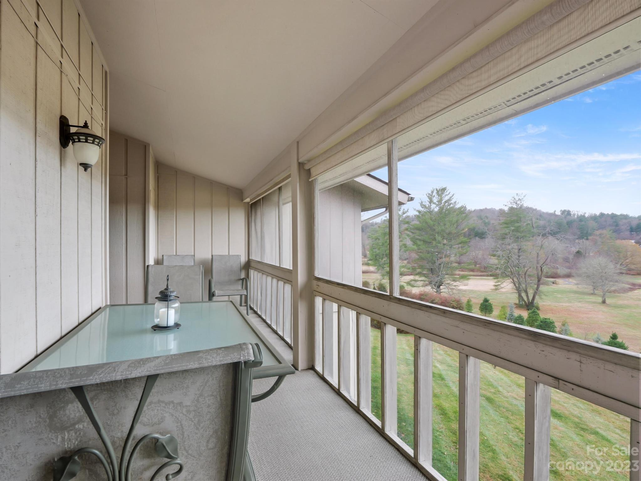 Enclosed balcony with a glass-top table and chairs overlooking landscaped grounds and trees.