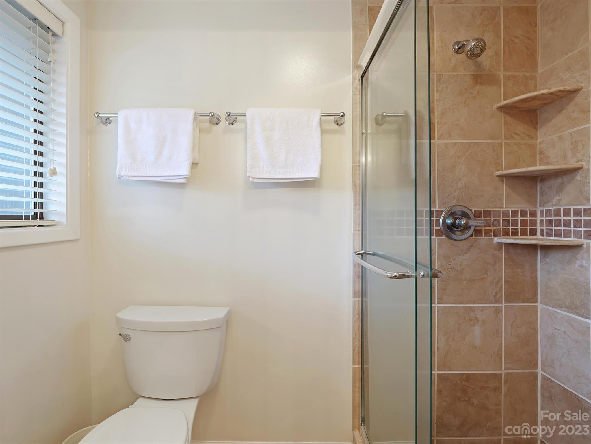 Bathroom with a glass shower, tan tile and built-in shelves beside a toilet and window.