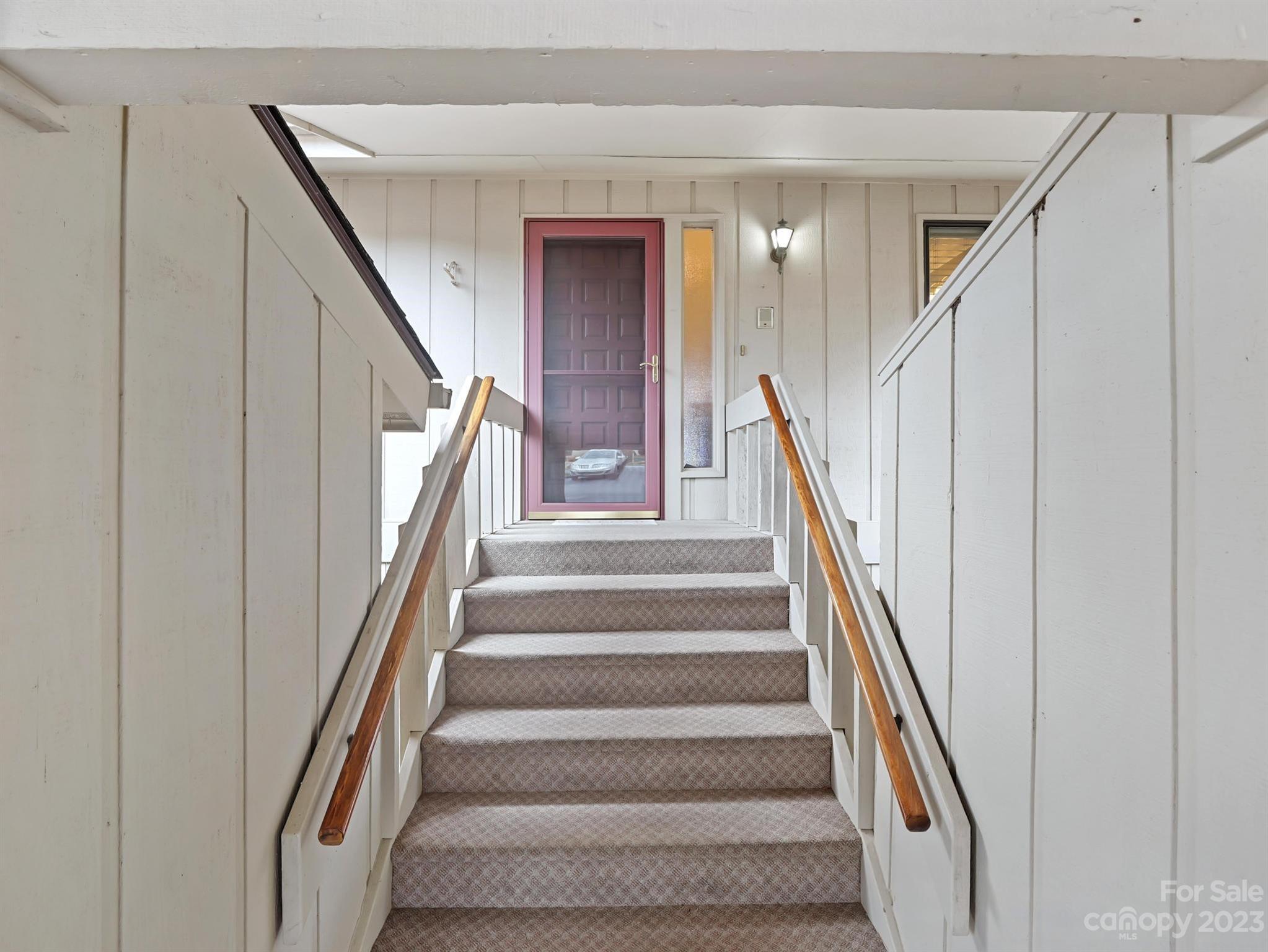 Carpeted staircase with wooden handrails leading to a burgundy entry door.